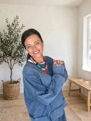 A woman smiling and posing indoors, wearing a blue denim jacket over a white top and a colorful beaded necklace. She is standing in a bright room with a potted plant, a wooden bench, and a window in the background.