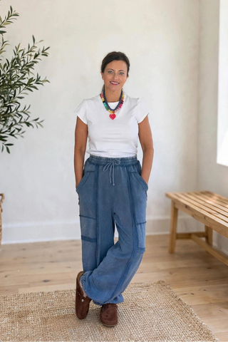 A woman stands in a bright, minimalist room wearing wide-leg blue cargo pants, a white t-shirt, and brown shoes. She is positioned in front of a large potted olive tree and a wooden bench.