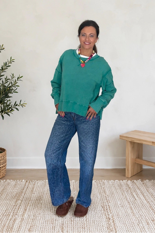 A woman stands in a bright room wearing a green crewneck sweater, blue jeans, and brown shoes, with a potted plant and a wooden bench in the background.