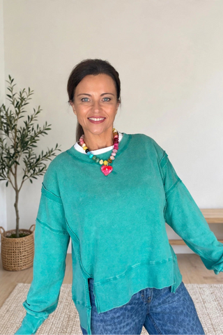 A woman wearing a teal-green long-sleeve top and blue jeans, standing in a brightly lit indoor room with a potted plant and wooden bench in the background.