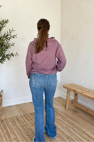 A woman viewed from behind wearing a dusty purple hoodie and blue jeans, standing in a minimalist room with cream walls, a potted olive tree, and a wooden bench.