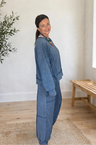 A woman wearing a blue denim jumpsuit stands in a bright, modern room with a potted olive tree, a wooden bench, and a woven rug.