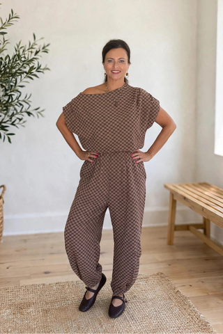 A woman modeling a brown and beige patterned off-the-shoulder jumpsuit in a bright, modern room with a potted olive tree and a wooden bench.