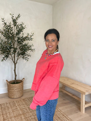A woman wearing a bright pink long-sleeve sweatshirt and blue jeans stands in a room with cream walls, a potted olive tree, and a wooden bench.
