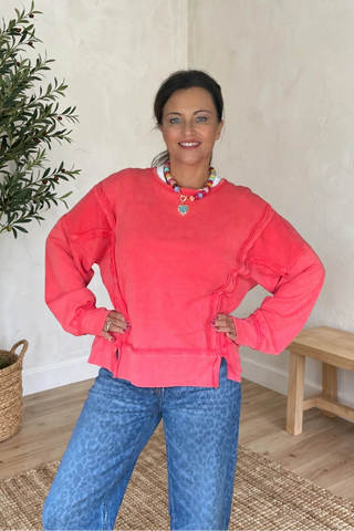 A woman wearing a coral pink long-sleeve sweatshirt and blue jeans stands in a room with textured cream walls, a potted olive tree, and a wooden bench.
