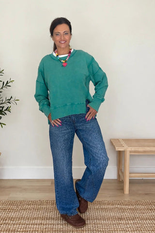 A woman wearing a teal green crewneck sweatshirt and blue jeans stands in a bright room with a potted plant, a woven rug, and a wooden bench.