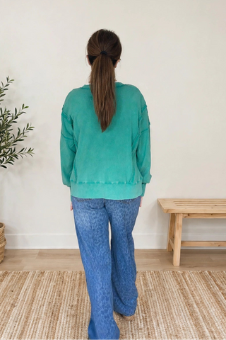 A woman viewed from behind wearing a teal-green long-sleeve knit top and blue jeans, standing on a woven rug in a bright room with a potted plant and a wooden bench.