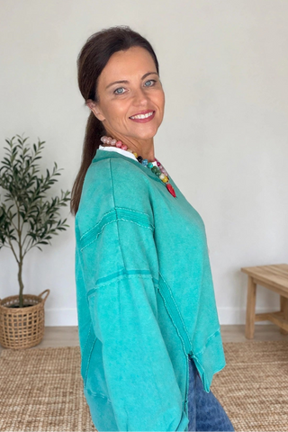 A woman wearing a turquoise long-sleeve knit top and blue jeans, standing in a brightly lit room with a potted plant and a wooden bench in the background.