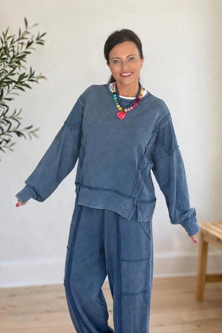 A woman modeling a matching two-piece lounge set in a bright, neutral room. The set consists of a long-sleeve cropped sweatshirt and matching wide-leg pants in a dusty slate blue color.