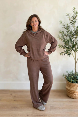 A woman modeling a matching brown lounge set consisting of a hooded sweatshirt and wide-leg sweatpants, standing in a brightly lit room with a textured beige wall, light wood floor, and a potted plant.