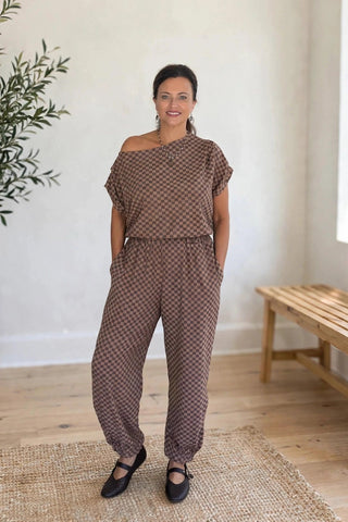 A woman modeling a brown and beige patterned off-the-shoulder jumpsuit in a bright, neutral room with a potted tree and a wooden bench.