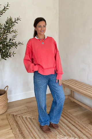A woman wearing a coral pink long-sleeve crewneck sweatshirt and blue jeans stands in a bright room with a potted plant, a wooden bench, and a woven rug.