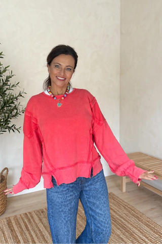 A woman wearing a coral pink long-sleeve top and blue jeans, standing in a brightly lit room with a potted plant and a woven bench in the background.