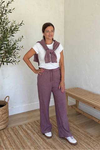 A woman modeling a dusty purple outfit consisting of wide-leg drawstring trousers and a matching knotted scarf, styled with a white t-shirt and white sneakers in a bright room with a potted olive tree and a wooden bench.