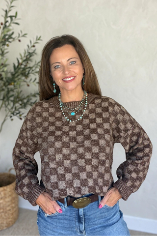 A woman wearing a brown and beige checkered knit sweater, blue jeans, and turquoise jewelry, standing in front of a light-colored wall next to a potted plant.