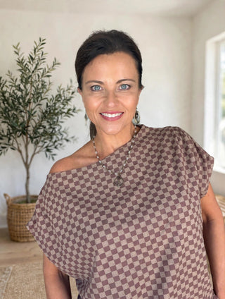A smiling woman with dark hair pulled back, wearing a brown and beige checkerboard patterned top with one shoulder, standing in a brightly lit indoor setting with a potted olive tree in the background.