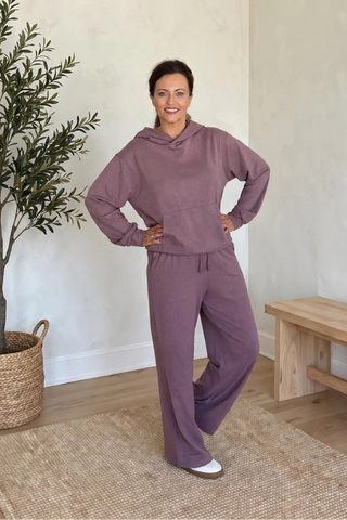 A woman modeling a matching dusty purple lounge set consisting of a long-sleeve hooded sweatshirt and wide-leg sweatpants, standing in a brightly lit room with a potted olive tree, a wooden bench, and a woven rug.