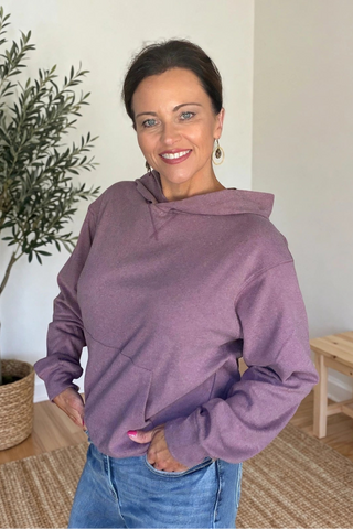 A woman wearing a dusty purple oversized hoodie and blue jeans, standing in a brightly lit room with a potted olive tree and a wooden bench in the background.