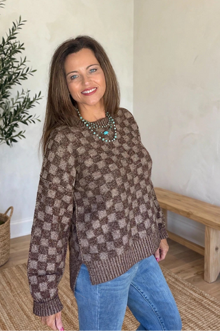 A woman wearing a brown and beige patterned knit sweater and blue jeans, standing in a room with a potted olive tree, a wooden bench, and a woven rug.