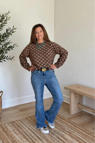 A woman stands in a bright room wearing a brown and beige patterned long-sleeve top, blue jeans, and white sneakers. A potted olive tree, a wooden bench, and a woven rug are visible in the background.