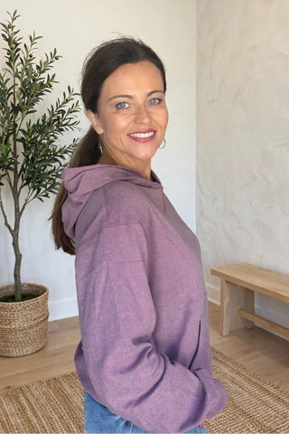 A woman wearing a heathered purple hoodie and blue jeans, smiling and looking back over her shoulder in a bright room with a potted plant and a wooden bench.