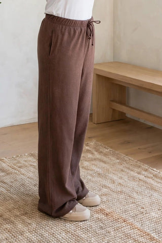 A woman stands in a bright, minimalist room with textured cream walls, a potted olive tree, and a wooden bench, wearing a white short-sleeve top and brown wide-leg drawstring pants.