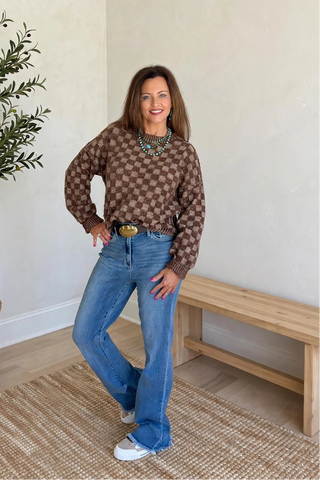 A woman modeling a brown and beige patterned knit sweater, blue jeans, and white sneakers in a bright room with a potted olive tree and a wooden bench.