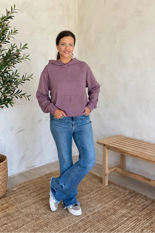 A woman stands in a bright room wearing a dusty purple pullover hoodie, blue jeans, and white sneakers, posing next to a potted olive tree and a wooden bench.