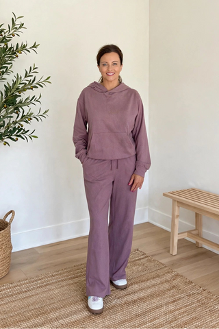 A woman modeling a matching dusty purple lounge set consisting of a cropped hoodie and wide-leg sweatpants, standing in a bright room with a potted plant and a wooden bench.