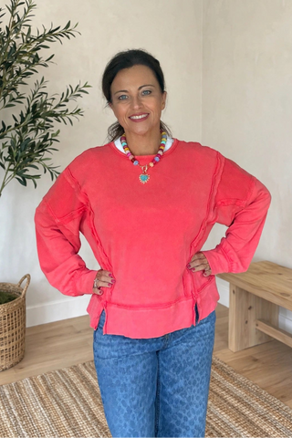 A woman wearing a coral pink long-sleeve top and blue jeans stands indoors with her hands on her hips, next to a potted olive tree and a wooden bench.