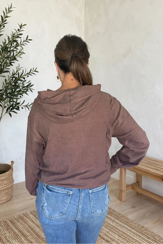 A woman viewed from behind wearing a dusty rose-brown cropped hoodie and blue jeans, standing in a brightly lit room with textured white walls, a potted olive tree, and a wooden bench.