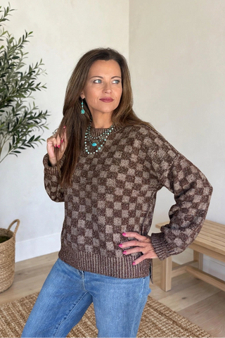 A woman wearing a brown and beige checkered knit sweater and blue jeans, posing in a brightly lit room with a potted olive tree and a wooden bench.
