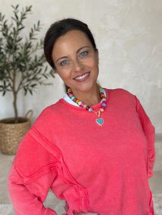 A smiling woman wearing a coral pink long-sleeve sweatshirt and a colorful beaded necklace with a heart-shaped pendant, standing in front of a textured light beige wall with a potted olive tree in the background.