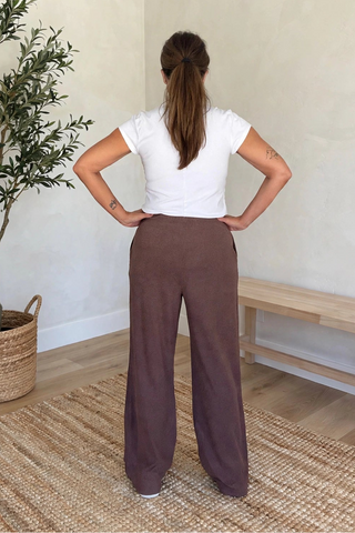 A woman stands with her back to the camera in a minimalist room, wearing loose brown wide-leg pants, a white t-shirt, and white sneakers. The room features a potted olive tree, a wooden bench, and a woven rug.