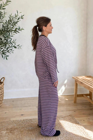 A woman stands in profile wearing a matching long-sleeve pajama set with thin horizontal purple and beige stripes, in a bright room with a potted olive tree, wooden bench, and window.