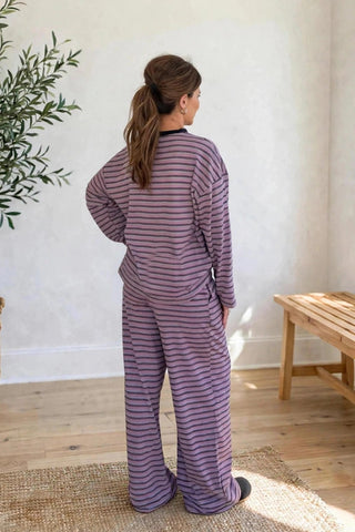 A woman stands with her back to the camera wearing a matching purple and pink striped long-sleeve pajama set in a bright, modern room with a potted olive tree and a wooden bench.