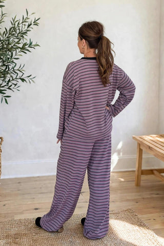 A woman stands with her back to the camera wearing a matching long-sleeve top and wide-leg pants set with horizontal purple and gray stripes. She is in a bright room with a potted olive tree, a wooden bench, and a window.