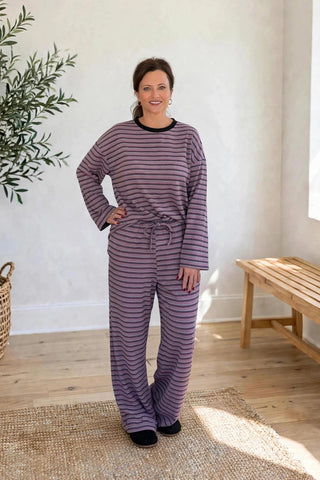 A woman modeling a matching two-piece pajama set with horizontal purple and gray stripes, featuring a long-sleeve top and matching pants, standing in a bright, modern room with a potted olive tree and a wooden bench.