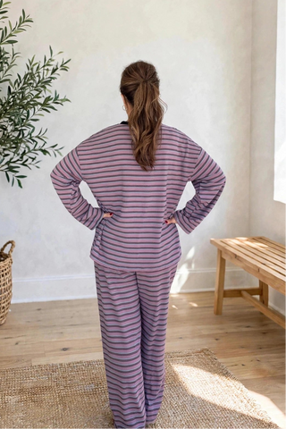 A woman stands with her back to the camera wearing a matching two-piece pajama set with horizontal stripes in dusty purple and white. She is photographed in a bright, modern room with a potted olive tree, a wooden bench, and a window.