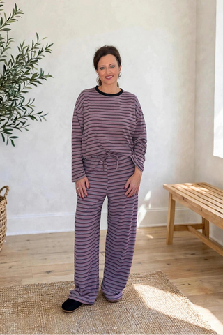 A woman wearing a matching two-piece pajama set with horizontal purple and gray stripes, standing in a bright room with a potted tree, a wooden bench, and a window.