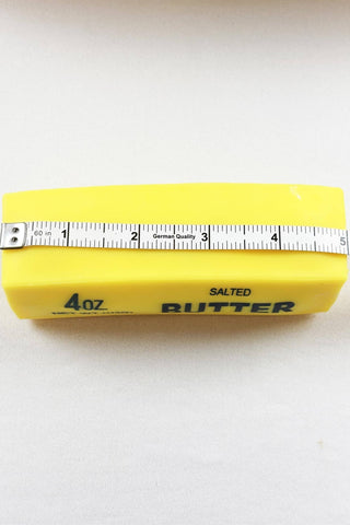 A yellow 4 oz stick of salted butter with a metal measuring tape ruler laid across the top, photographed on a plain light background.