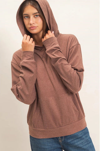 A woman wearing a heathered brown pullover hoodie with a front kangaroo pocket and drawstrings, paired with blue jeans, set against a plain light beige background.