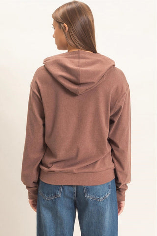 A woman viewed from the back wearing a dusty rose-brown pullover hoodie and blue jeans against a plain off-white studio background.