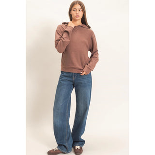 A woman modeling a brown long-sleeve hooded sweatshirt with wide-leg blue jeans and brown loafers, standing against a plain off-white studio background.