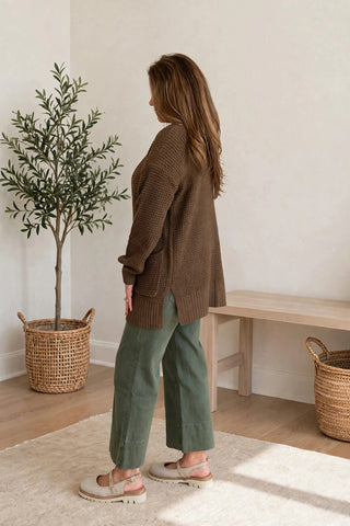 A woman stands in a bright, minimalist room wearing a brown ribbed knit cardigan, sage green wide-leg pants, and beige sandals. She is shown from the back, with a potted olive tree, a wooden bench, and woven baskets in the background.
