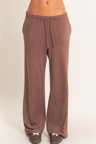 A person wearing loose, wide-leg lounge pants in a muted brown or taupe color, paired with dark slip-on shoes, shown against a plain light background.
