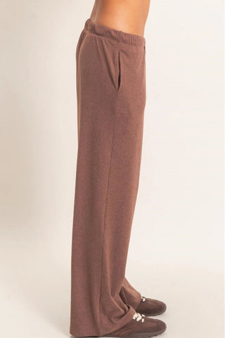 A person shown from the waist down wearing loose, straight-leg brown knit pants and brown loafers against a plain light beige studio background.