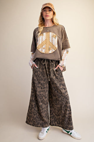 A model wearing a brown graphic t-shirt with a peace sign design, wide-leg leopard-print pants, a beige cap, and white sneakers, standing against a solid beige background.
