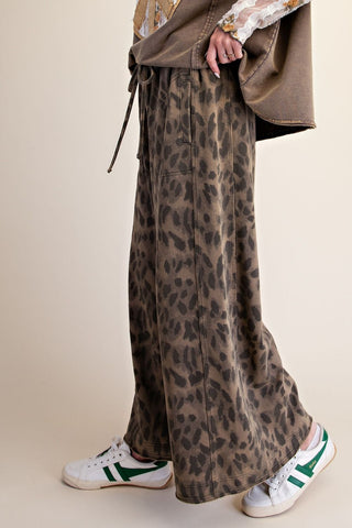 A person wearing wide-leg leopard-print pants in brown and beige tones, styled with a brown jacket and white sneakers with green stripes, against a plain beige studio background.