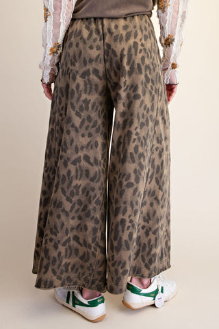 Wide-leg trousers in a brown and beige leopard print pattern, shown on a model from the back wearing white sneakers with green accents.